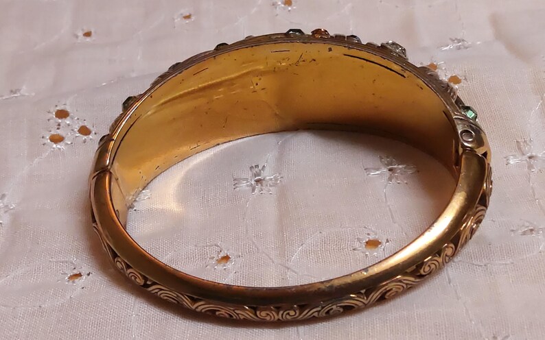 May include: A vintage gold-toned hinged bangle bracelet. The bracelet features an ornate, engraved design along the edges and is adorned with small, colorful gemstones. The interior has a warm, aged appearance, suggesting its antique nature.