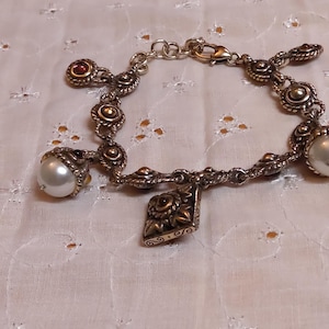 May include: Silver-toned bracelet with ornate detailing and multiple charms. Features large faux pearls and small red-toned accents. A decorative charm with a floral design hangs from the center of the bracelet. The bracelet is displayed on a white, patterned surface.