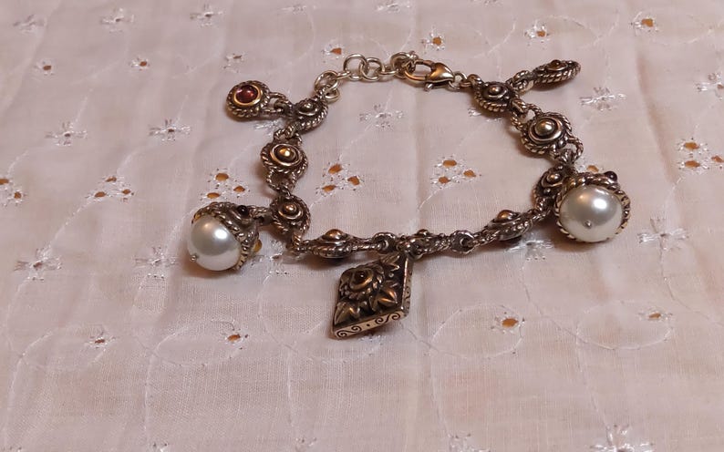 May include: Silver-toned bracelet with ornate detailing and multiple charms. Features large faux pearls and small red-toned accents. A decorative charm with a floral design hangs from the center of the bracelet. The bracelet is displayed on a white, patterned surface.