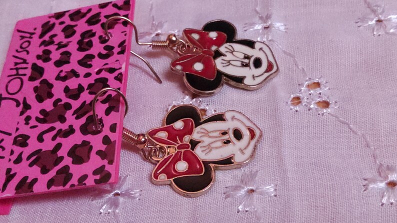 Pu&ograve; includere: Un paio di orecchini pendenti di Minnie Mouse. Ogni orecchino presenta un viso bianco con dettagli neri, un fiocco rosso a pois bianchi e orecchie nere. Gli orecchini sono dorati e hanno una chiusura a gancio.