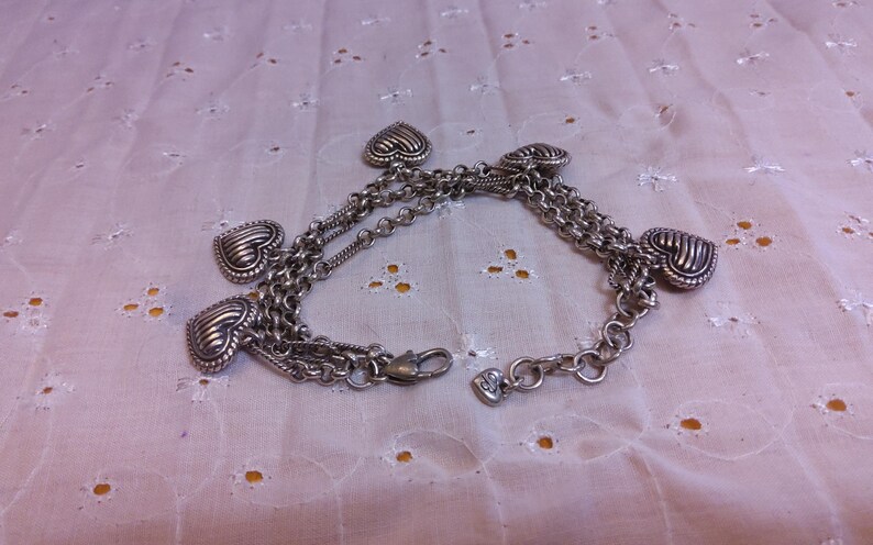 Pu&ograve; includere: Braccialetto in argento con pi&ugrave; ciondoli a forma di cuore. Il braccialetto &egrave; composto da pi&ugrave; catene e ha una chiusura a moschettone.