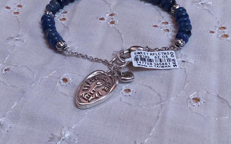 Puede incluir: Una pulsera de cuentas azules oscuras con detalles plateados. Un dije plateado con las palabras "LOVE THE" est&aacute; adjunto, junto con un peque&ntilde;o dije de coraz&oacute;n. Tambi&eacute;n se adjunta una etiqueta con texto.