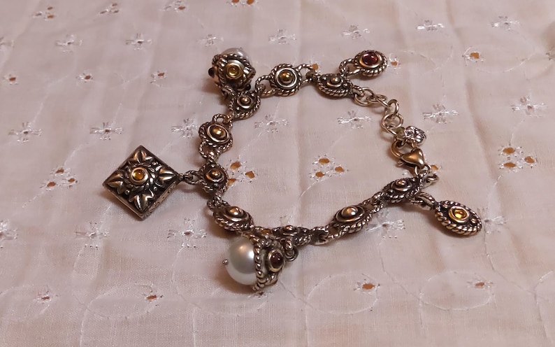 May include: Silver-toned charm bracelet with ornate detailing. Features a square charm, round charms with gold accents, and a dangling pearl. The bracelet has a lobster clasp closure and is set against a white, textured background.