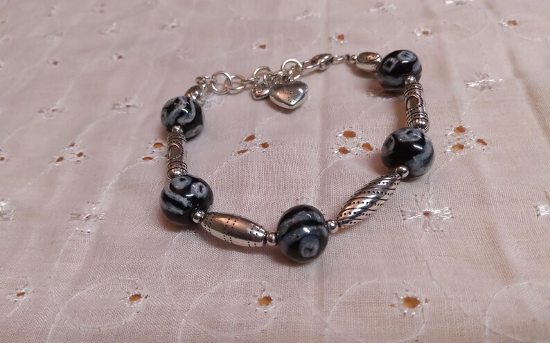 May include: A beaded bracelet featuring black and white patterned beads interspersed with silver-toned metal beads and accents. The bracelet includes a small heart charm and is displayed on a white, textured surface.