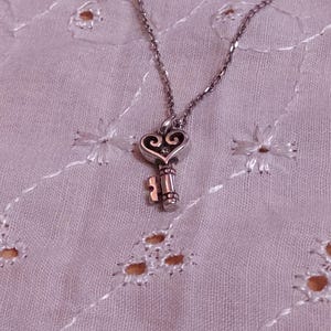 Op de afbeelding: Een zilverkleurige ketting met een sleutelhanger. De sleutel heeft een hartvormige bovenkant met decoratieve krullen en een gedetailleerde schacht. De ketting ligt op een lichtpaarse, geborduurde stof.