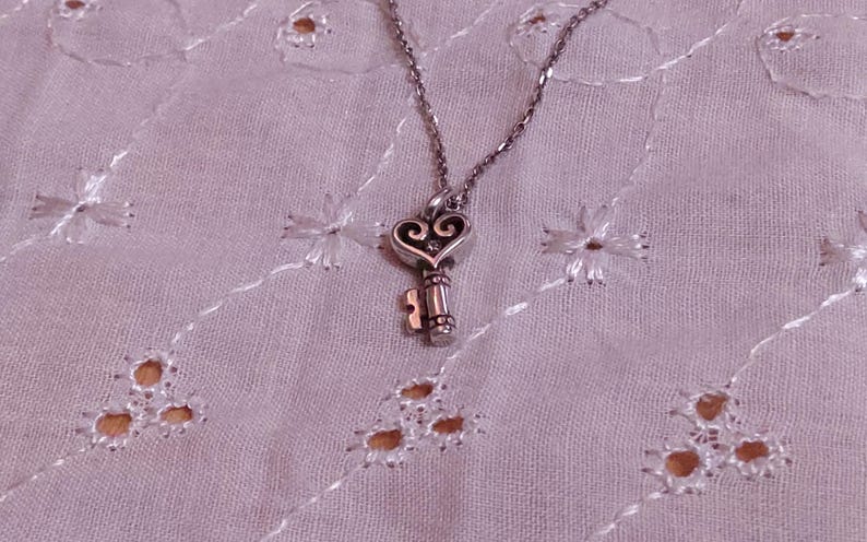 Op de afbeelding: Een zilverkleurige ketting met een sleutelhanger. De sleutel heeft een hartvormige bovenkant met decoratieve krullen en een gedetailleerde schacht. De ketting ligt op een lichtpaarse, geborduurde stof.