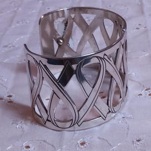 May include: A silver cuff bracelet with an openwork design. The bracelet features a series of interconnected ribbon-like shapes. The metal has a polished finish, reflecting light. The bracelet is a wide style, suitable for a statement piece.