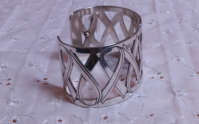 May include: A silver cuff bracelet with an openwork design. The bracelet features a series of interconnected ribbon-like shapes. The metal has a polished finish, reflecting light. The bracelet is a wide style, suitable for a statement piece.