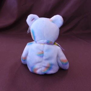 May include: A light blue plush bear with a rainbow pattern. The bear has a yellow ribbon around its neck.