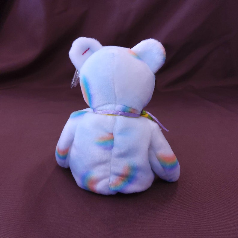 May include: A light blue plush bear with a rainbow pattern. The bear has a yellow ribbon around its neck.