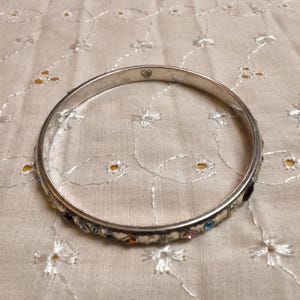May include: A silver bangle bracelet with small, colorful gemstones. The bracelet has a heart-shaped design on the top. The bracelet is resting on a cream-colored, embroidered fabric.