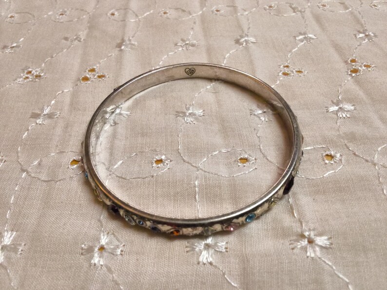 May include: A silver bangle bracelet with small, colorful gemstones. The bracelet has a heart-shaped design on the top. The bracelet is resting on a cream-colored, embroidered fabric.