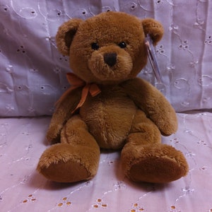 May include: A brown teddy bear with a light orange bow sitting on a white crocheted surface.