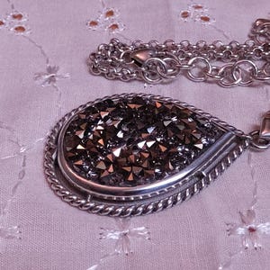 Może przedstawiać: Srebrny naszyjnik z dużą zawieszką w kształcie łzy. Zawieszka wypełniona jest małymi, ciemnoszarymi kryształkami i ma skręconą, sznurowatą obw&oacute;dkę. Łańcuszek naszyjnika jest r&oacute;wnież srebrny. Dołączona jest biała metka z czarnym tekstem.