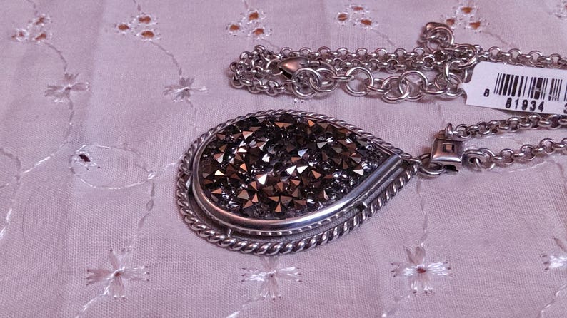 Może przedstawiać: Srebrny naszyjnik z dużą zawieszką w kształcie łzy. Zawieszka wypełniona jest małymi, ciemnoszarymi kryształkami i ma skręconą, sznurowatą obw&oacute;dkę. Łańcuszek naszyjnika jest r&oacute;wnież srebrny. Dołączona jest biała metka z czarnym tekstem.