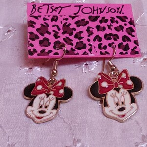 Pu&ograve; includere: Un paio di orecchini di Minnie Mouse di Betsey Johnson. Ogni orecchino presenta un contorno dorato, un viso bianco, orecchie nere e un fiocco rosso a pois bianchi. Gli orecchini pendono da un gancio dorato.