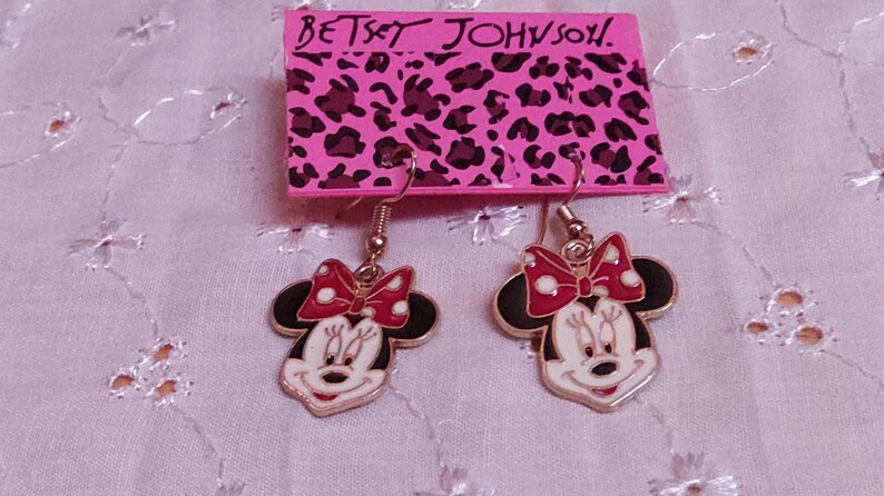 Pu&ograve; includere: Un paio di orecchini di Minnie Mouse di Betsey Johnson. Ogni orecchino presenta un contorno dorato, un viso bianco, orecchie nere e un fiocco rosso a pois bianchi. Gli orecchini pendono da un gancio dorato.
