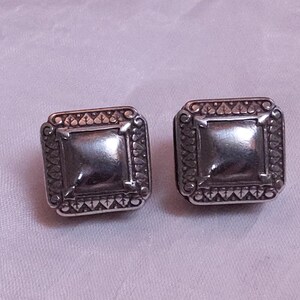 May include: A pair of square silver-toned stud earrings. Each earring features a central square design surrounded by an ornate, textured border. The earrings are displayed on a light purple surface.