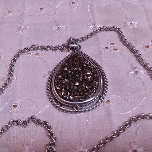 Może przedstawiać: Srebrny naszyjnik z wisiorkiem w kształcie łzy. Wisiorek wypełniony jest małymi, ciemnymi kryształkami i ma ozdobną srebrną obw&oacute;dkę. Łańcuszek jest r&oacute;wnież srebrny i ma klasyczny wz&oacute;r ogniw.