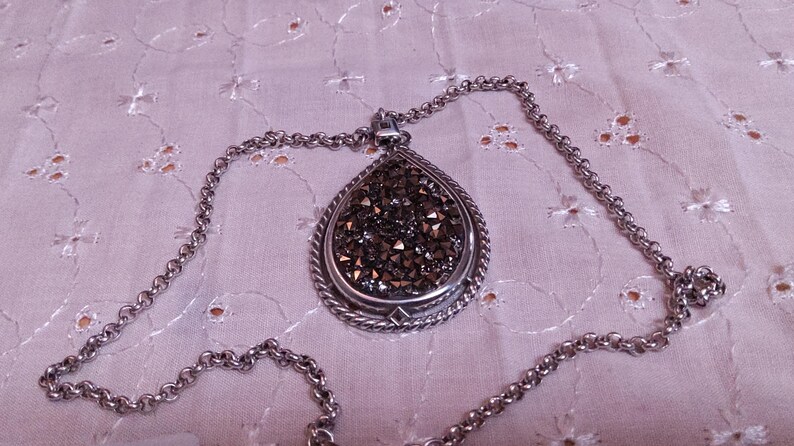 Może przedstawiać: Srebrny naszyjnik z wisiorkiem w kształcie łzy. Wisiorek wypełniony jest małymi, ciemnymi kryształkami i ma ozdobną srebrną obw&oacute;dkę. Łańcuszek jest r&oacute;wnież srebrny i ma klasyczny wz&oacute;r ogniw.