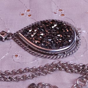 Może przedstawiać: Srebrny naszyjnik z wisiorkiem w kształcie łzy wypełnionym ciemnymi, błyszczącymi kryształami. Wisiorek jest obramowany skręconym sznurkiem. Łańcuszek jest w klasycznym stylu ogniw z zapięciem na karabińczyk.