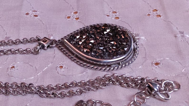 Może przedstawiać: Srebrny naszyjnik z wisiorkiem w kształcie łzy wypełnionym ciemnymi, błyszczącymi kryształami. Wisiorek jest obramowany skręconym sznurkiem. Łańcuszek jest w klasycznym stylu ogniw z zapięciem na karabińczyk.