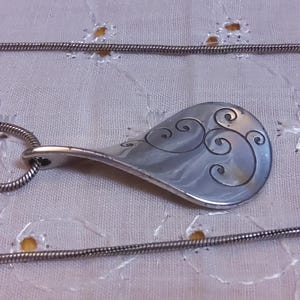 May include: A silver necklace with a pendant shaped like a teardrop with a swirling design. The chain is a thin silver snake chain.
