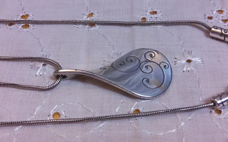 May include: A silver necklace with a pendant shaped like a teardrop with a swirling design. The chain is a thin silver snake chain.