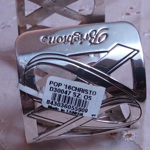 May include: Silver-toned metal napkin ring with a ribbon design. The word "Brighton" is embossed on the top. A white tag with text and a barcode is attached to the napkin ring. The napkin ring is on a light pink surface.