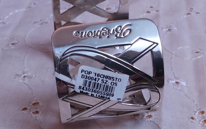 May include: Silver-toned metal napkin ring with a ribbon design. The word "Brighton" is embossed on the top. A white tag with text and a barcode is attached to the napkin ring. The napkin ring is on a light pink surface.