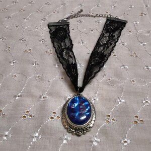 May include: A black lace choker necklace featuring a central oval pendant with a polished blue iridescent cabochon set in a silver-toned metal frame with decorative filigree details and a thin metal chain extender at the top.