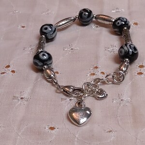 May include: A silver bracelet with black and white patterned beads and a heart charm. The bracelet features a mix of round and oval beads, with decorative silver accents. The heart charm hangs from a clasp.