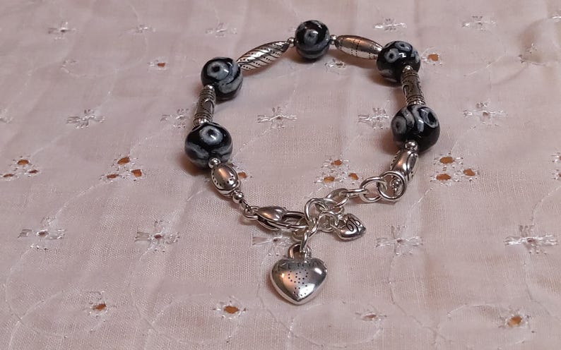 May include: A silver bracelet with black and white patterned beads and a heart charm. The bracelet features a mix of round and oval beads, with decorative silver accents. The heart charm hangs from a clasp.