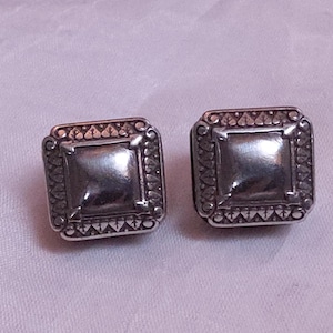 May include: A pair of square silver-toned stud earrings. Each earring features a raised square center and a decorative border with a repeating pattern. The earrings are displayed on a light purple fabric.