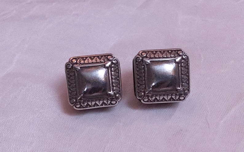 May include: A pair of square silver-toned stud earrings. Each earring features a raised square center and a decorative border with a repeating pattern. The earrings are displayed on a light purple fabric.