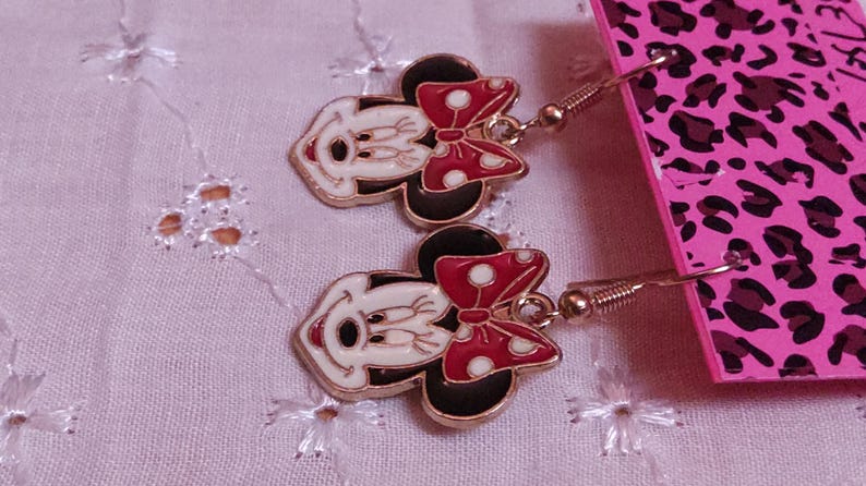 Pu&ograve; includere: Un paio di orecchini dorati di Minnie Mouse. Ogni orecchino presenta un viso bianco, orecchie nere e un fiocco rosso a pois bianchi. Gli orecchini sono su uno sfondo rosa.