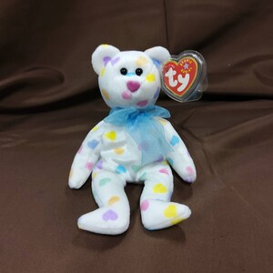 May include: White plush beanie baby bear with a blue ribbon around its neck. The bear is covered in colorful heart-shaped spots. The bear has a Ty tag attached to its ear.