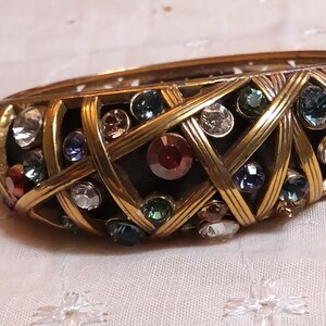 May include: A vintage gold-tone bangle bracelet with a woven design, encrusted with colorful rhinestones. The bracelet features a variety of jewel-toned stones, including red, blue, green, and clear, set within the gold metalwork.