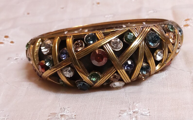 May include: A vintage gold-tone bangle bracelet with a woven design, encrusted with colorful rhinestones. The bracelet features a variety of jewel-toned stones, including red, blue, green, and clear, set within the gold metalwork.
