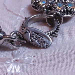 May include: A silver charm with a flower design and a tag that reads 'Alexas Angel'. The charm is attached to a silver chain.
