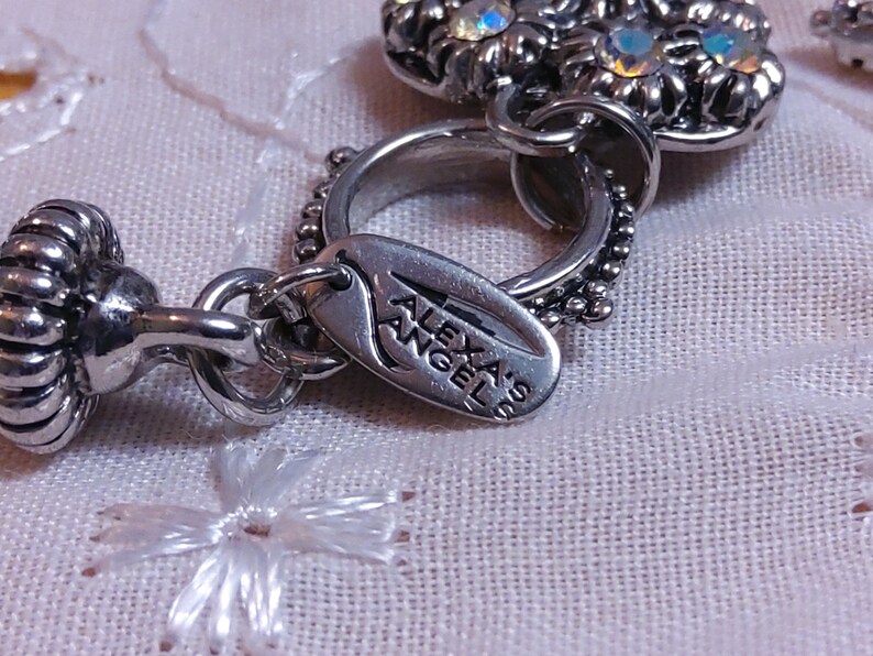 May include: A silver charm with a flower design and a tag that reads 'Alexas Angel'. The charm is attached to a silver chain.