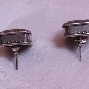 May include: A pair of silver-toned stud earrings. Each earring has a rectangular shape with a textured top and small, decorative rivets along the bottom edge. The earrings are on a light purple background.