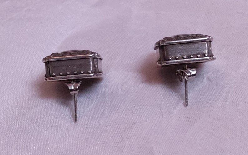 May include: A pair of silver-toned stud earrings. Each earring has a rectangular shape with a textured top and small, decorative rivets along the bottom edge. The earrings are on a light purple background.