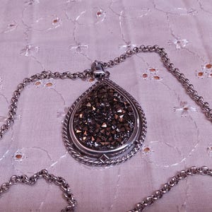 Może przedstawiać: Srebrny naszyjnik z wisiorkiem w kształcie łzy. Wisiorek wypełniony jest ciemnymi, błyszczącymi kryształami i obramowany skręconym sznurkiem. Łańcuszek ma ogniwowy wz&oacute;r.