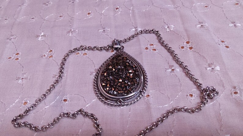 Może przedstawiać: Srebrny naszyjnik z wisiorkiem w kształcie łzy. Wisiorek wypełniony jest ciemnymi, błyszczącymi kryształami i obramowany skręconym sznurkiem. Łańcuszek ma ogniwowy wz&oacute;r.