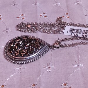 Może przedstawiać: Srebrny naszyjnik z wisiorkiem w kształcie łzy wypełnionym ciemnymi, błyszczącymi kryształami. Wisiorek jest obramowany skręconym sznurkiem. Łańcuszek jest w delikatnym stylu ogniw. Do łańcuszka przymocowana jest metka z kodem kreskowym.