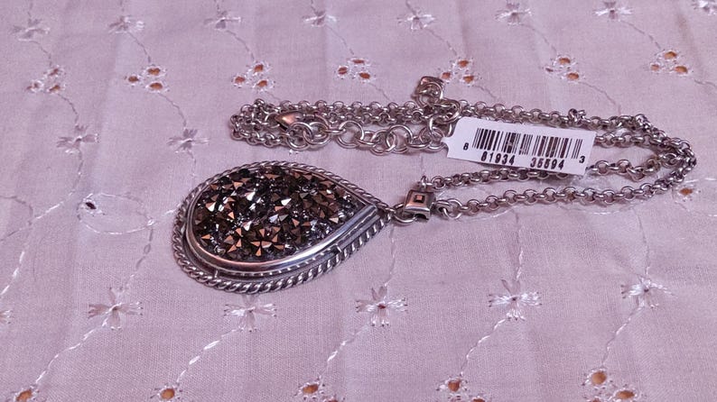 Może przedstawiać: Srebrny naszyjnik z wisiorkiem w kształcie łzy wypełnionym ciemnymi, błyszczącymi kryształami. Wisiorek jest obramowany skręconym sznurkiem. Łańcuszek jest w delikatnym stylu ogniw. Do łańcuszka przymocowana jest metka z kodem kreskowym.