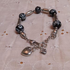 May include: A silver bracelet featuring black and white patterned beads and decorative silver accents. The bracelet includes a heart-shaped charm and a smaller charm with the initials "BB".