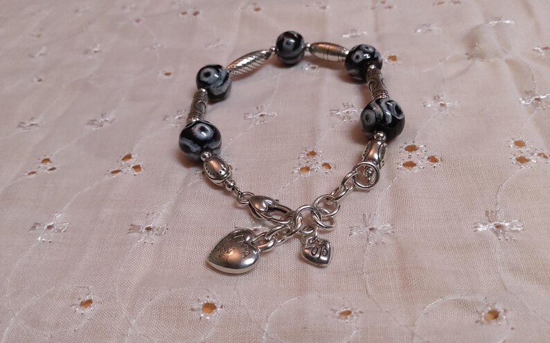 May include: A silver bracelet featuring black and white patterned beads and decorative silver accents. The bracelet includes a heart-shaped charm and a smaller charm with the initials "BB".