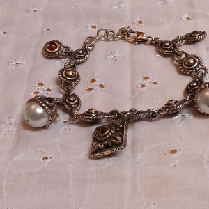 May include: Silver-toned bracelet with ornate detailing, featuring pearl and red gemstone accents. The bracelet has a decorative charm and is displayed on a white, patterned surface. The jewelry is a classic accessory.